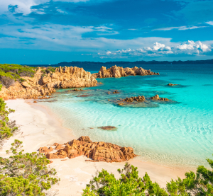 Traumhafter Sandstrand mit türkisblauem Wasser und rötlichen Felsen auf Sardinien, umgeben von grüner Mittelmeer-Vegetation unter blauem Himmel mit Wolken.