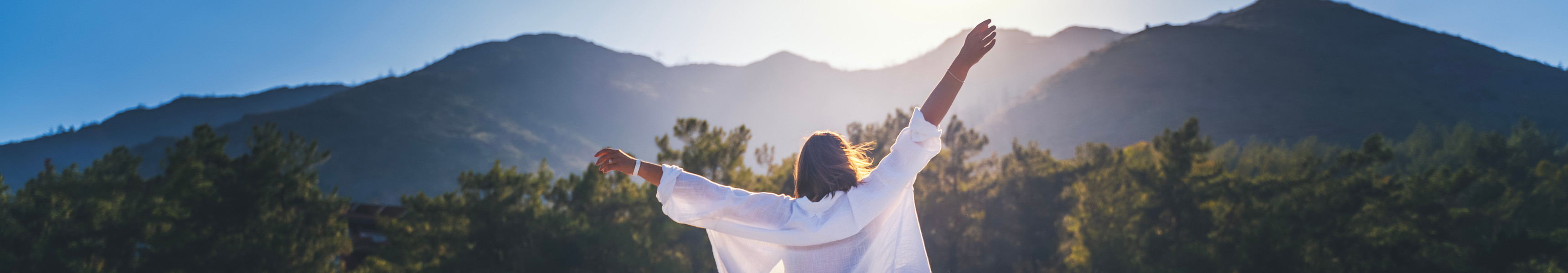 Eine Frau in einem weißen Hemd genießt mit erhobenen Händen die Morgensonne vor der Kulisse der Berge bei Sonnenaufgang