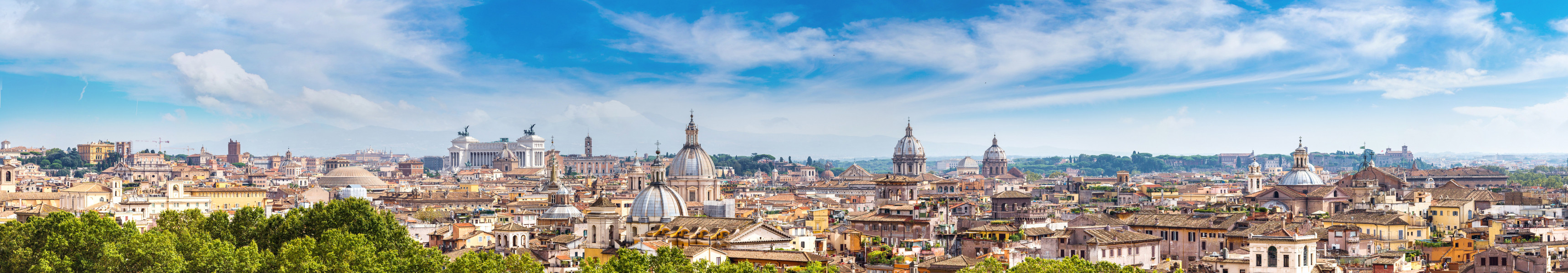 Panoramablick auf die Dächer von Rom, mit markanten historischen Gebäuden, Kirchen und Kuppeln, unter einem klaren blauen Himmel.