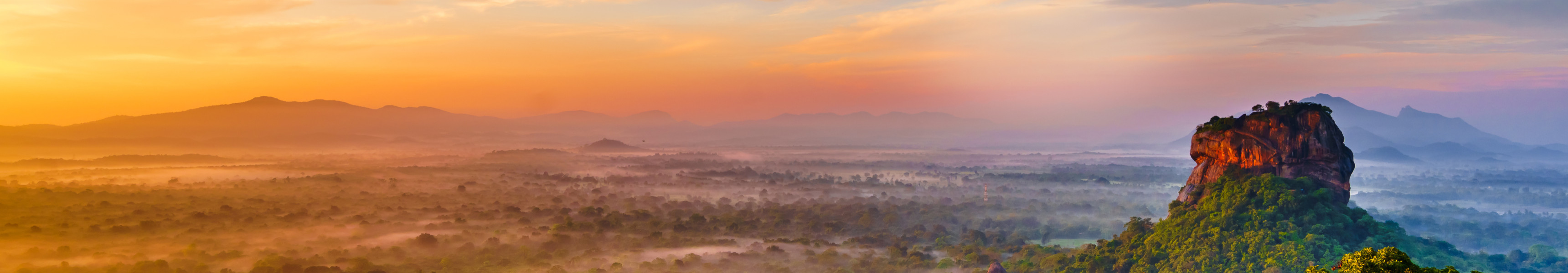 Luftaufnahme des Sigiriya-Felsens in Sri Lanka bei Sonnenaufgang mit grünem Dschungel im Vordergrund und farbenprächtigem Himmel im Hintergrund