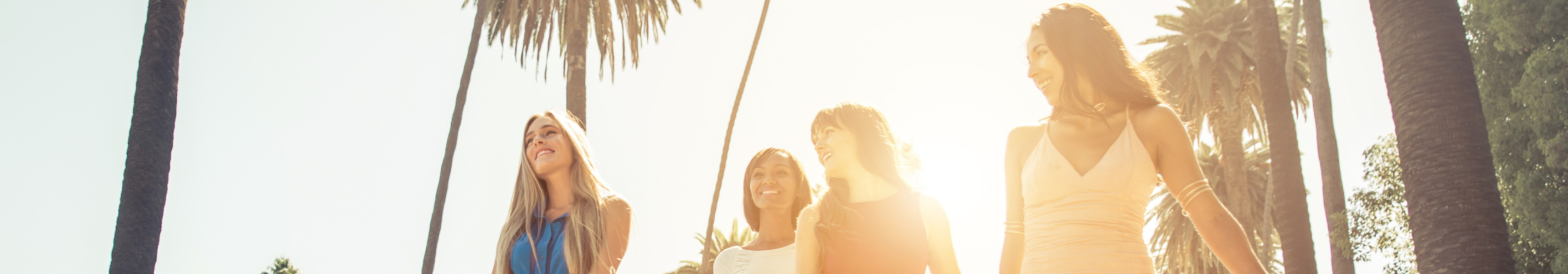 Vier junge Frauen in bunten Sommerkleidern gehen lachend mit Einkaufst&uuml;ten eine von Palmen ges&auml;umte Stra&szlig;e entlang, w&auml;hrend die Sonne im Hintergrund hell scheint.