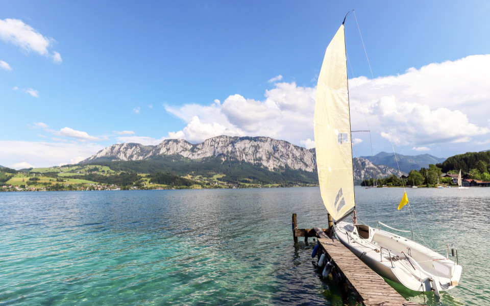 Ein Segelboot liegt an einem Steg am Attersee im Salzkammergut, umgeben von gr&uuml;nen H&uuml;geln und Bergen, unter einem klaren, blauen Himmel.