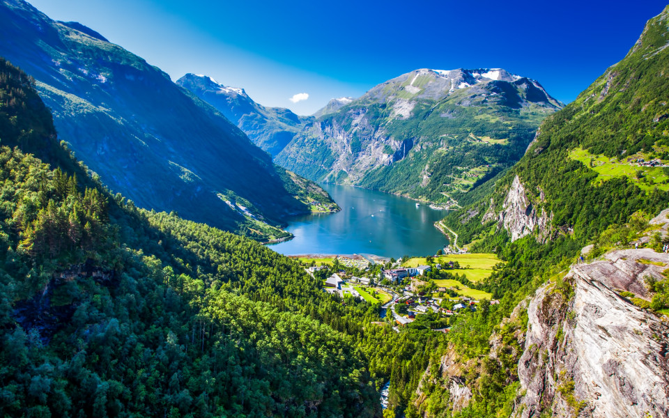 Der Geirangerfjord erstreckt sich tief zwischen steilen, grünen Berghängen mit schneebedeckten Gipfeln und einem kleinen Dorf am Ufer.