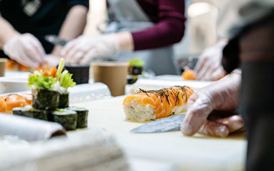 Mehrere Personen bereiten an einer Theke Sushi zu; im Vordergrund liegt eine frisch gerollte Lachsrolle mit Reis und Nori, daneben weitere Sushi-St&uuml;cke und Zutaten auf einem hellen Schneidebrett.